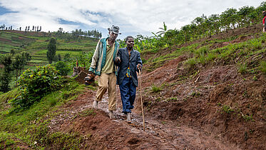 The image depicts two individuals navigating a rough, muddy terrain, likely in a rural area. One person appears to be using a walking stick, suggesting mobility may be challenging in this environment. The setting is natural and green, with cultivated lands in the background, indicating an agricultural landscape.